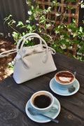 White handbag on a wooden table with two cups of coffee and saucers.