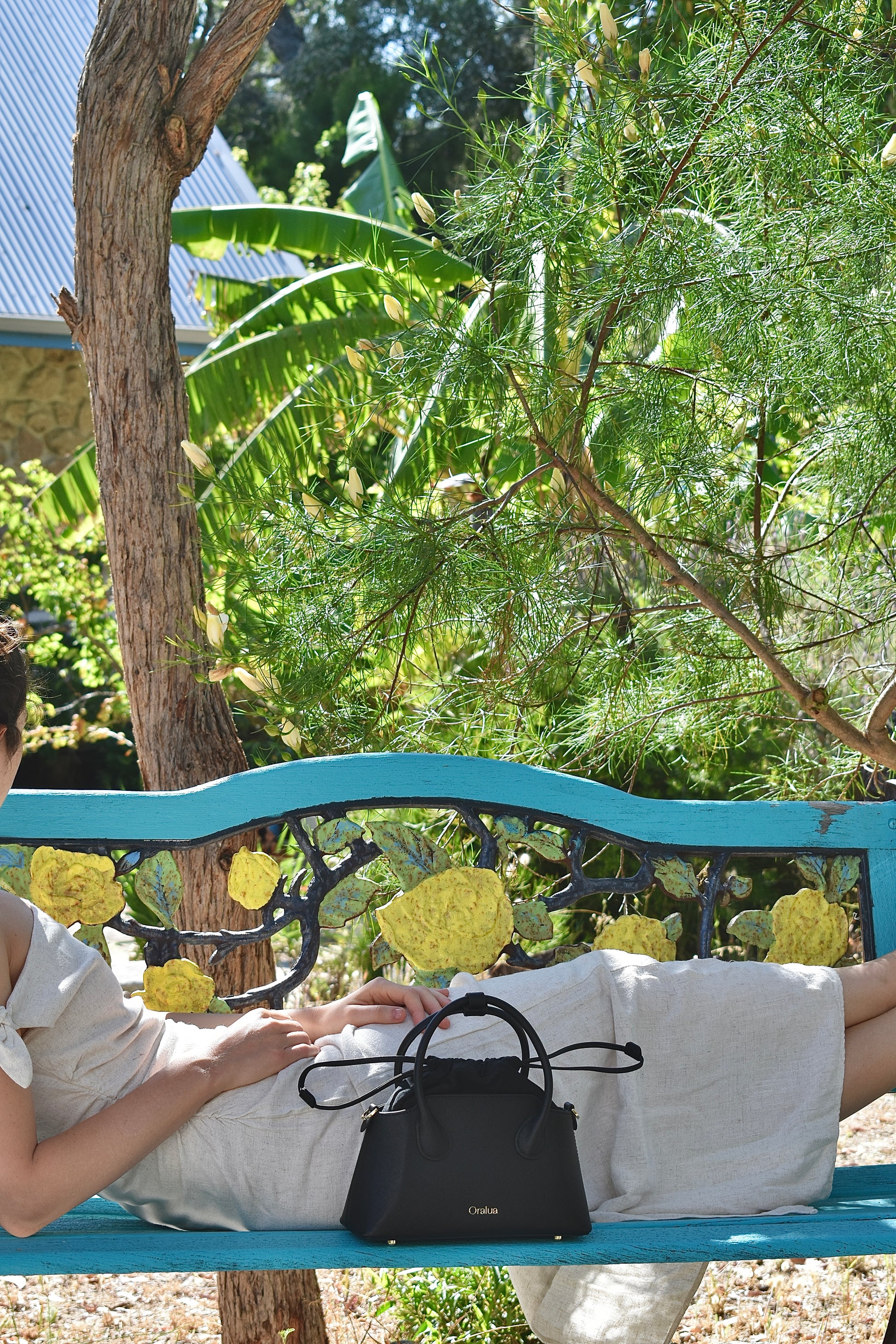 Person sitting on a blue bench with a black handbag in a garden setting
