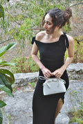 Woman in a black dress holding a white handbag outdoors with greenery in the background